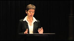 A woman in a black coat and white shirt is giving a lecture. Her hands are animated, and she is standing in front of a black curtain.