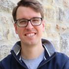 A man in glasses faces the viewer and smiles. He is wearing a navy blue jumper and pale blue t shirt.